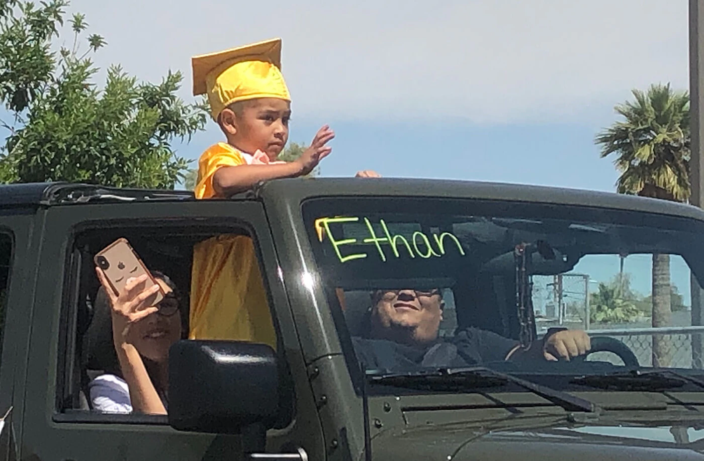 child in graduation cap and gown waves from a car's sunroof