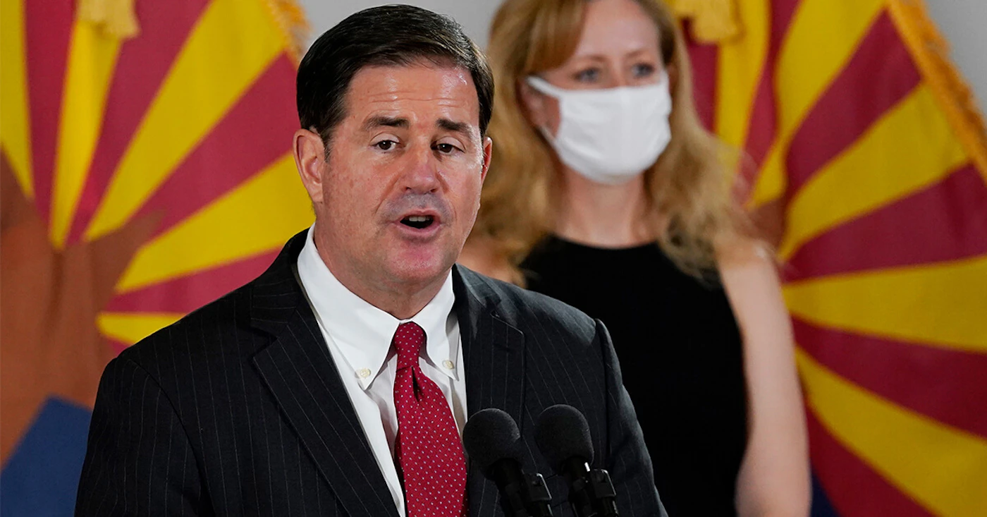 Gov. Ducey without mask standing in front of Dr. Cara Christ with masks, both in front of Arizona state flags