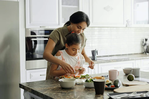woman standing behind young girl as they prepare food