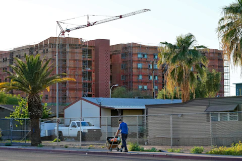 A man selling fruit walks past the fenced-off Periwinkle Mobile Home Park, Thursday, April 11, 2023, in Phoenix as new student housing is constructed in the background. Residents of the park are facing an eviction deadline of May 28 due to a private university's plan to redevelop the land for student housing. (AP Photo/Matt York)