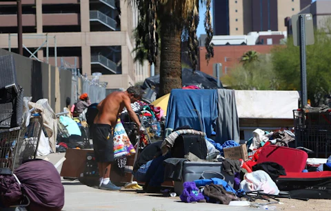 FILE - A man sweeps the sidewalk as tents and crude lean-tos crowd the sidewalks where many homeless people live along the streets on March 24, 2020, in Phoenix. The city is wrangling with two dueling lawsuits as it tries to manage a homelessness crisis that has converted its downtown into a tent city housing hundreds of people under the blazing desert sun just as summer temperatures soar into the 90s. (AP Photo/Ross D. Franklin, File)