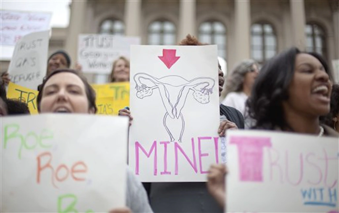 women holding protest signs in front of Georgia capital; one is a drawing of a uterus that says mine