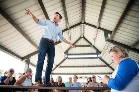 Jeff Jackson, in a blue button-down shirt with his sleeves rolled up, stands on a picnic table at a town hall addressing attendees in Gastonia, NC.