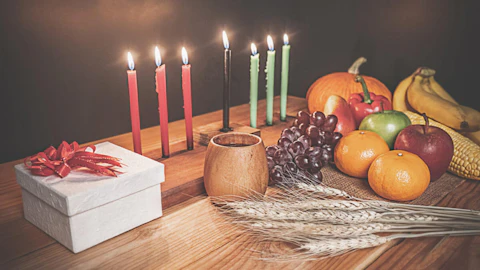 A traditional Kwanzaa table adorned with a kinara, umoja cup, fruits, and a gift. Image by Shutterstock.